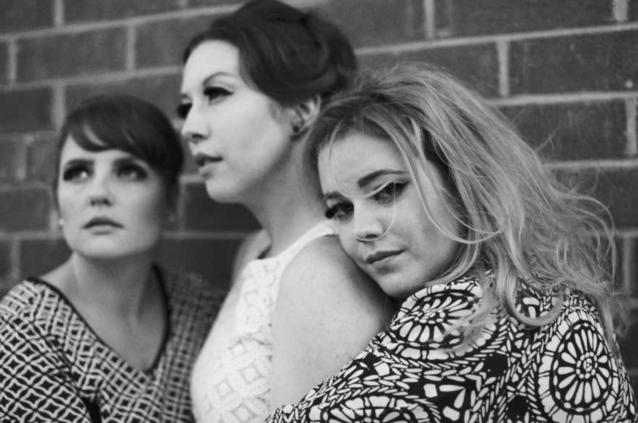 Alex, Maggie and Lizzy in a black and white photograph with hair and makeup in the style of a 50s girl group. The 3 women are hugging each other.